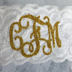 Pode incluir: Jarreteira de renda branca com um monograma de glitter dourado. O monograma est&aacute; em uma fonte de escrita e apresenta as letras "CJM".