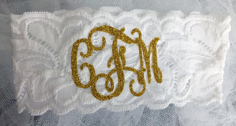 Pode incluir: Jarreteira de renda branca com um monograma de glitter dourado. O monograma est&aacute; em uma fonte de escrita e apresenta as letras "CJM".