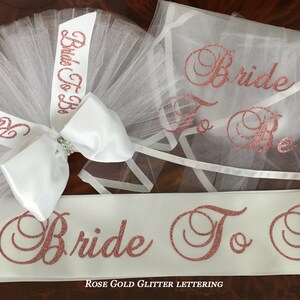 Pode incluir: Uma faixa de noiva branca com letras de glitter dourado rosa que dizem "Bride To Be". A faixa &eacute; combinada com um v&eacute;u de tule branco com um la&ccedil;o de cetim branco e um adorno de pedraria. O v&eacute;u tamb&eacute;m tem letras de glitter dourado rosa que dizem "Bride To Be".