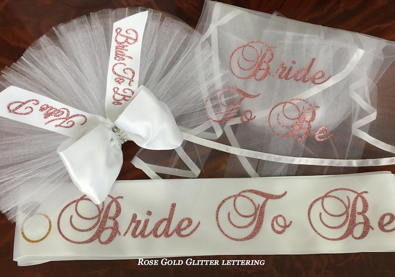 Pode incluir: Uma faixa de noiva branca com letras de glitter dourado rosa que dizem "Bride To Be". A faixa &eacute; combinada com um v&eacute;u de tule branco com um la&ccedil;o de cetim branco e um adorno de pedraria. O v&eacute;u tamb&eacute;m tem letras de glitter dourado rosa que dizem "Bride To Be".