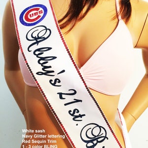 May include: White satin sash with navy blue glitter lettering that reads "Abby's 21st. Birth". The sash is trimmed with red sequins and features a blue and red Chicago Cubs logo.