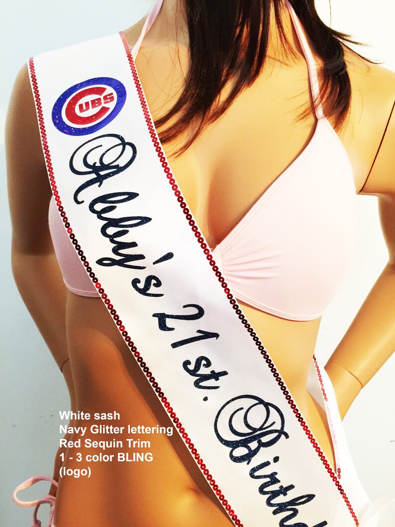 May include: White satin sash with navy blue glitter lettering that reads "Abby's 21st. Birth". The sash is trimmed with red sequins and features a blue and red Chicago Cubs logo.