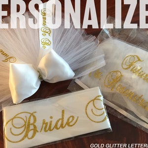 Pode incluir: Um v&eacute;u de tule branco com um la&ccedil;o de cetim branco e uma fita de brilho dourado que diz "June 6, 2018" e "Here To Be". Tamb&eacute;m est&aacute; inclu&iacute;da uma faixa branca com letras de brilho dourado que diz "Bride" e uma bolsa de tule branco com letras de brilho dourado que diz "Future Mrs. [Last Name]". O texto "GOLD GLITTER LETTERING" est&aacute; na parte inferior da imagem.