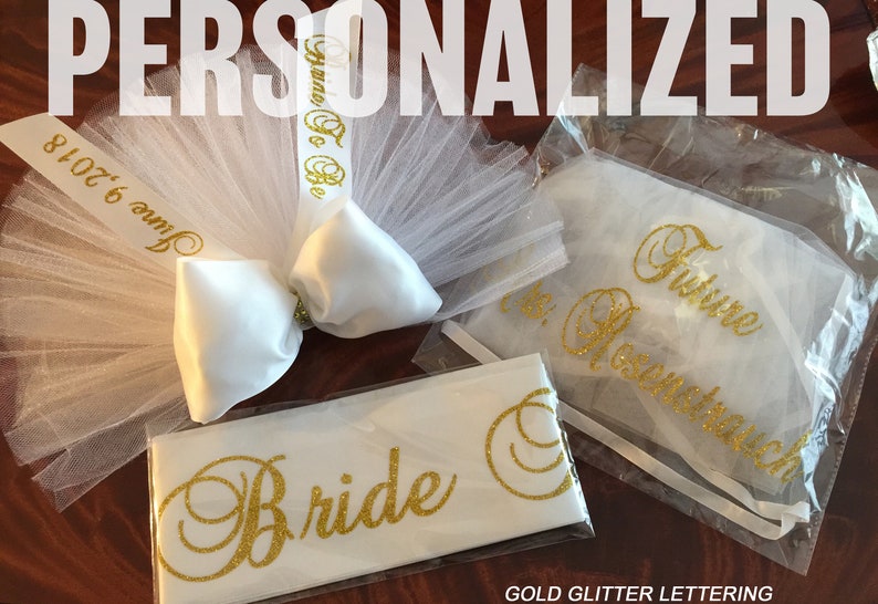 Pode incluir: Um v&eacute;u de tule branco com um la&ccedil;o de cetim branco e uma fita de brilho dourado que diz "June 6, 2018" e "Here To Be". Tamb&eacute;m est&aacute; inclu&iacute;da uma faixa branca com letras de brilho dourado que diz "Bride" e uma bolsa de tule branco com letras de brilho dourado que diz "Future Mrs. [Last Name]". O texto "GOLD GLITTER LETTERING" est&aacute; na parte inferior da imagem.