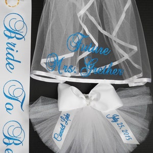 Pode incluir: Faixa de noiva branca com letras de glitter azul que diz "Bride To Be". Um v&eacute;u branco com letras de glitter azul que diz "Future Mrs. Grether". Um la&ccedil;o de tule branco com letras de glitter azul que diz "Married Life" e "July 25, 2015".