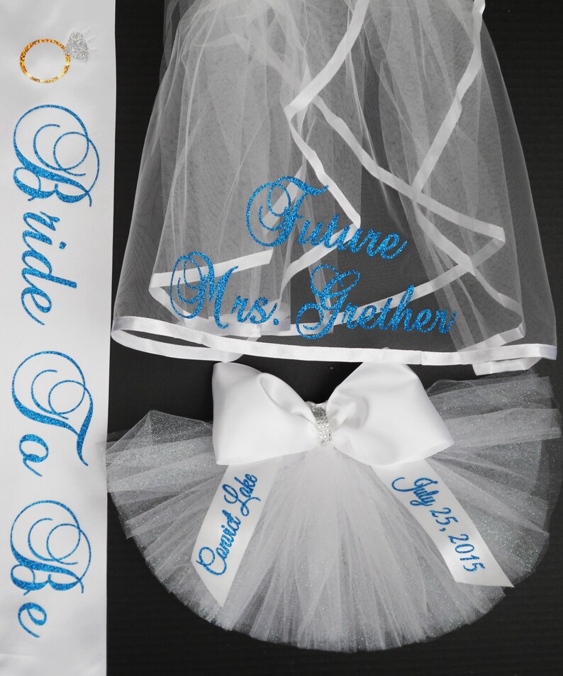 Pode incluir: Faixa de noiva branca com letras de glitter azul que diz "Bride To Be". Um v&eacute;u branco com letras de glitter azul que diz "Future Mrs. Grether". Um la&ccedil;o de tule branco com letras de glitter azul que diz "Married Life" e "July 25, 2015".