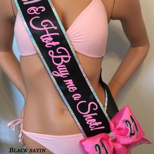 K&ouml;nnte beinhalten: Schwarzer Satinschleife mit silbernem Besatz und pinkfarbenem Schriftzug "21 & Hot Buy me a Shot!"  Die Schleife hat eine gro&szlig;e rosa Schleife mit zwei silbernen "21"-Zahlen.