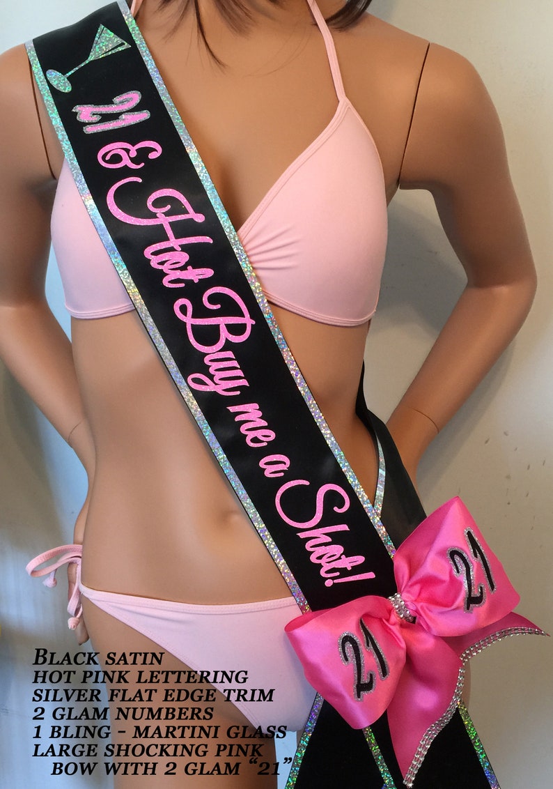 K&ouml;nnte beinhalten: Schwarzer Satinschleife mit silbernem Besatz und pinkfarbenem Schriftzug "21 & Hot Buy me a Shot!"  Die Schleife hat eine gro&szlig;e rosa Schleife mit zwei silbernen "21"-Zahlen.
