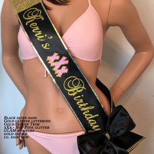 May include: Black satin sash with gold glitter lettering, gold sequin trim, and hot pink glitter "xxx" for a birthday celebration. The sash has a large black bow with gold trim.
