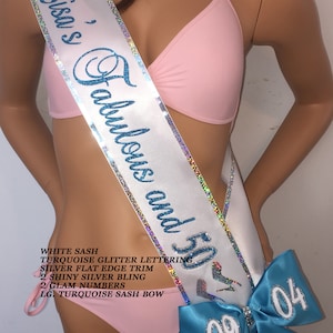 K&ouml;nnte beinhalten: Wei&szlig;er Satinsash mit t&uuml;rkisfarbenem Glitzer-Schriftzug "Lisa's Fabulous and 50". Der Sash hat einen silbernen, flachen Rand und zwei gl&auml;nzende silberne Verzierungen. Eine gro&szlig;e t&uuml;rkisfarbene Schleife mit silbernen Glitzerzahlen "09 04" ist am Sash befestigt.