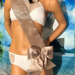 May include: A sparkly gold sash with a large satin bow. The bow is decorated with the name "Angela" and the number "30".