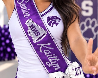 May include: A white and purple cheerleading uniform featuring a sash with the text Senior, a megaphone graphic with HHS, the name Kaittyn, and a large white bow with the text Wildcats and 27, accented with silver glitter detailing.