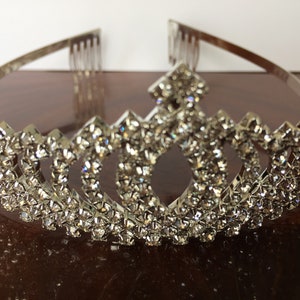 Pode incluir: Tiara de prata com design de coroa, adornada com uma fileira de strass brilhantes.