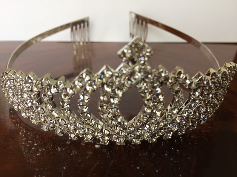 Pode incluir: Tiara de prata com design de coroa, adornada com uma fileira de strass brilhantes.