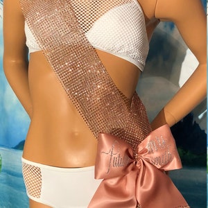 May include: A rose gold rhinestone sash with a satin bow that says "Future Mrs. Smith". The sash is draped over a white bikini top and bottom. The bow is embellished with rhinestones.