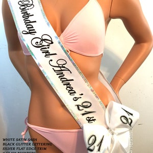 K&ouml;nnte beinhalten: Wei&szlig;er Satin-Sch&auml;rpe mit schwarzer Glitzer-Beschriftung, silbernem flachem Rand, zwei gro&szlig;en, gl&auml;nzenden Zahlen und einer gro&szlig;en Schleife. Die Sch&auml;rpe tr&auml;gt die Aufschrift "Birthday Girl Andrea's 21st".