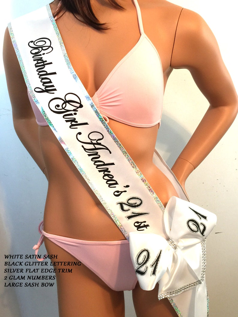 K&ouml;nnte beinhalten: Wei&szlig;er Satin-Sch&auml;rpe mit schwarzer Glitzer-Beschriftung, silbernem flachem Rand, zwei gro&szlig;en, gl&auml;nzenden Zahlen und einer gro&szlig;en Schleife. Die Sch&auml;rpe tr&auml;gt die Aufschrift "Birthday Girl Andrea's 21st".