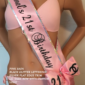 K&ouml;nnte beinhalten: Rosa Satin-Sch&auml;rpe mit silbernem Besatz und schwarzer Glitzer-Beschriftung "Chanel's 21st Birthday". Eine gro&szlig;e rosa Schleife mit einem schwarzen "C" und der Zahl "21" ist an der Sch&auml;rpe befestigt.