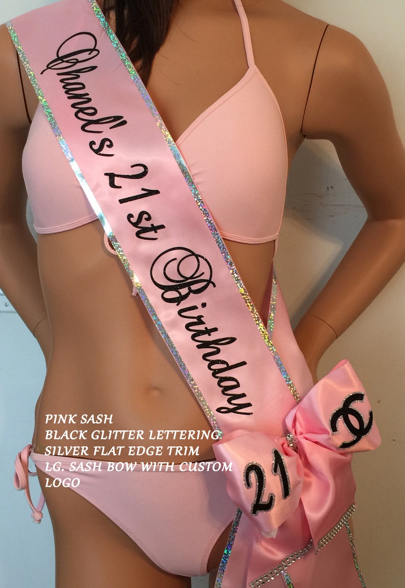 K&ouml;nnte beinhalten: Rosa Satin-Sch&auml;rpe mit silbernem Besatz und schwarzer Glitzer-Beschriftung "Chanel's 21st Birthday". Eine gro&szlig;e rosa Schleife mit einem schwarzen "C" und der Zahl "21" ist an der Sch&auml;rpe befestigt.