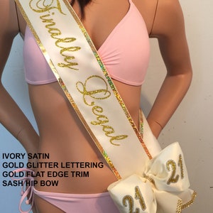 K&ouml;nnte beinhalten: Ein Sash aus elfenbeinfarbenem Satin mit goldenen Glitzerbuchstaben, auf dem "Finally Legal" steht, und einem goldenen, flachen Rand. Der Sash hat eine gro&szlig;e elfenbeinfarbene Schleife mit goldenen Glitzerbuchstaben, auf der auf jeder Seite "21" steht.