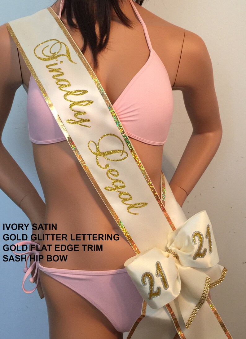 K&ouml;nnte beinhalten: Ein Sash aus elfenbeinfarbenem Satin mit goldenen Glitzerbuchstaben, auf dem "Finally Legal" steht, und einem goldenen, flachen Rand. Der Sash hat eine gro&szlig;e elfenbeinfarbene Schleife mit goldenen Glitzerbuchstaben, auf der auf jeder Seite "21" steht.