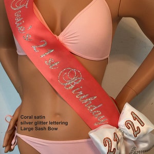 Puede incluir: Sash de sat&eacute;n coral con letras de purpurina plateada que dicen "21st Birthday". Un gran lazo de sat&eacute;n blanco con letras de purpurina plateada que dicen "21" est&aacute; unido al sash.