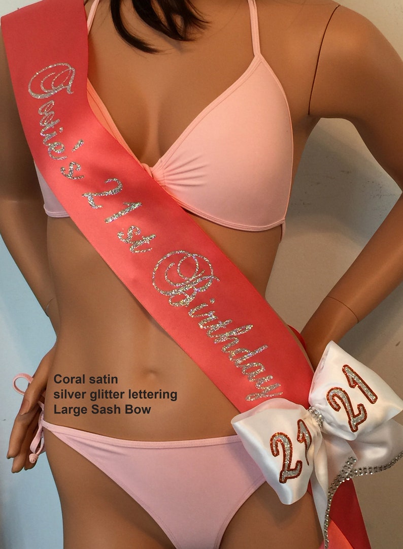 Puede incluir: Sash de sat&eacute;n coral con letras de purpurina plateada que dicen "21st Birthday". Un gran lazo de sat&eacute;n blanco con letras de purpurina plateada que dicen "21" est&aacute; unido al sash.