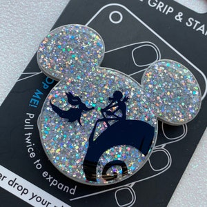 May include: A white glitter phone grip with a black silhouette of Jack Skellington and Sally from The Nightmare Before Christmas. The grip is shaped like Mickey Mouse ears and says "Pop Me! Pull twice to expand" on the back.