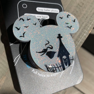 May include: A white glitter phone grip with a black silhouette of Jack Skellington from The Nightmare Before Christmas. The silhouette shows Jack standing in front of his spooky house with a fence and bats flying in the air. The phone grip is shaped like Mickey Mouse ears.