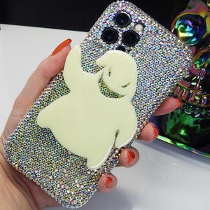 May include: A clear phone case with a sparkly, iridescent finish. A white ghost character is featured on the case. The case has a black camera lens with three lenses.