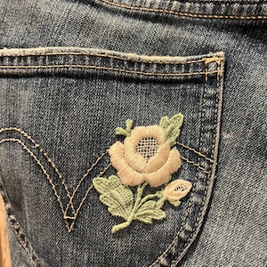May include: A pair of blue denim jeans with a white and green embroidered flower on the back pocket.