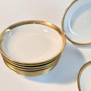 May include: A stack of eight white porcelain bowls with gold trim. The bowls are round and shallow, with a slightly flared rim.