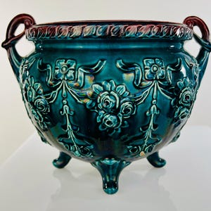 May include: A teal blue ceramic planter with three legs and two handles. The planter is decorated with raised floral designs and has a burgundy rim.