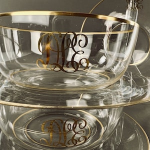 May include: A set of three clear glass bowls with gold rims. The bowls are stacked on top of each other. The top bowl has a gold monogram of the letters "D.E.G." on the inside. The bottom bowl has the same monogram on the outside.