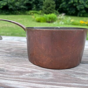 May include: A copper measuring cup with a single handle. The cup has a worn, aged look and is sitting on a wooden surface.