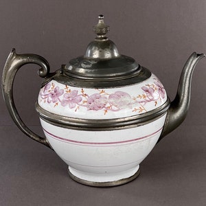May include: A vintage silver teapot with a white porcelain body decorated with pink floral designs. The teapot has a lid and a spout.