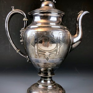 May include: A silver teapot with an ornate design and a grapevine motif on the lid. The teapot has a round body with a flared base and a spout on the right side. The teapot is engraved with the date "1892".