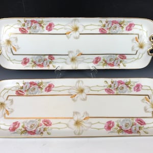 May include: Two rectangular serving trays with white backgrounds, adorned with floral patterns of white lilies and pink roses. The trays have gold trim and handles, creating an elegant aesthetic. The trays are likely for serving food or displaying items.