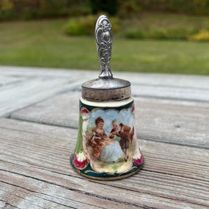 May include: A small porcelain bell with a silver top and handle. The bell is decorated with a painted scene of a couple in a garden setting.