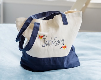 May include: A white canvas tote bag with blue trim and the name "Jackson" embroidered in black cursive. The bag has two small red and yellow airplanes embroidered on either side of the name.