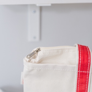 May include: A cream-colored canvas tote bag featuring a prominent vertical red stripe along the side. The bag includes a top zipper closure with a silver-toned metal pull tab and a small white fabric tag attached to the side seam near the zipper.