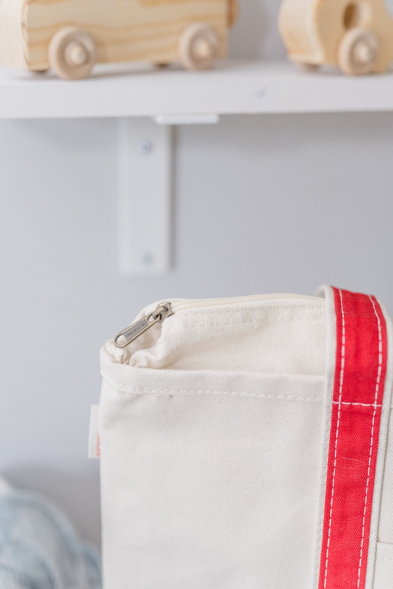 May include: A cream-colored canvas tote bag featuring a prominent vertical red stripe along the side. The bag includes a top zipper closure with a silver-toned metal pull tab and a small white fabric tag attached to the side seam near the zipper.