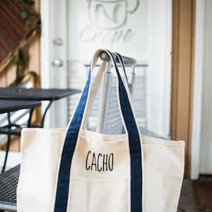 May include: A cream-colored canvas tote bag with navy blue trim and handles. The bag features the name CACHO embroidered in black capital letters on the front pocket. The design includes a reinforced bottom panel and two long shoulder straps.