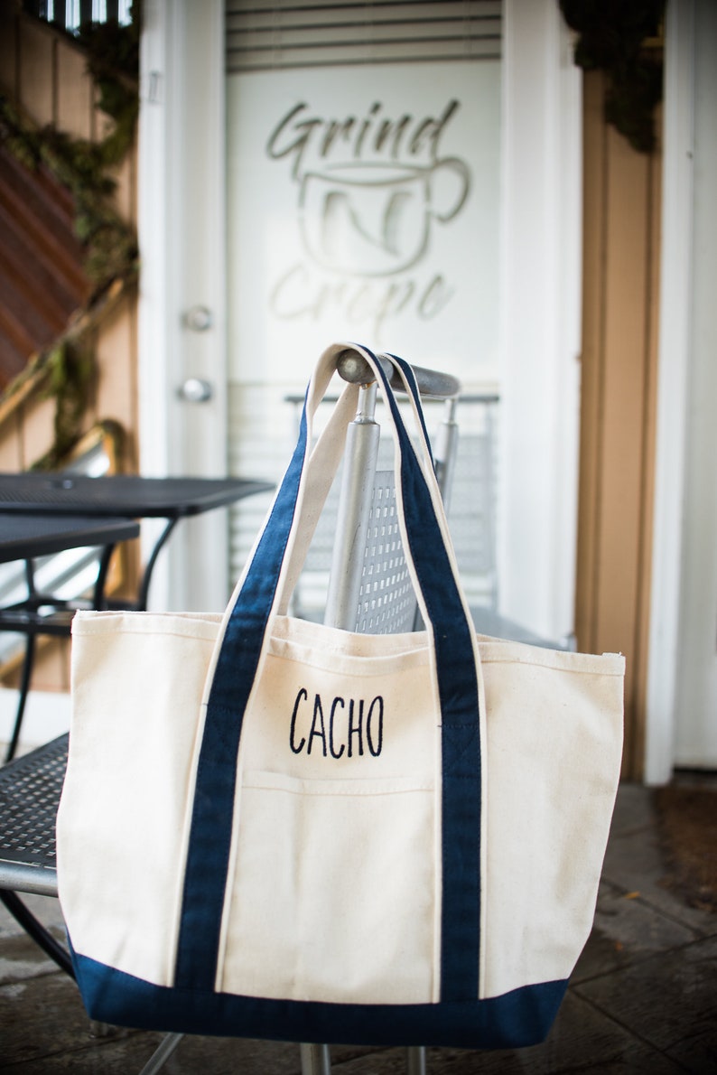 May include: A cream-colored canvas tote bag with navy blue trim and handles. The bag features the name CACHO embroidered in black capital letters on the front pocket. The design includes a reinforced bottom panel and two long shoulder straps.