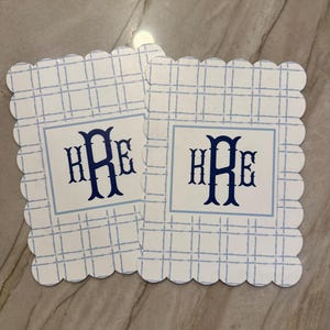 K&ouml;nnte beinhalten: Zwei quadratische Untersetzer mit gewelltem Rand. Jeder Untersetzer hat ein blau-wei&szlig;es Karomuster und ein zentrales Quadrat mit dem Monogramm "HRE" in Marineblau. Die Untersetzer sind aus einem robusten Material gefertigt.