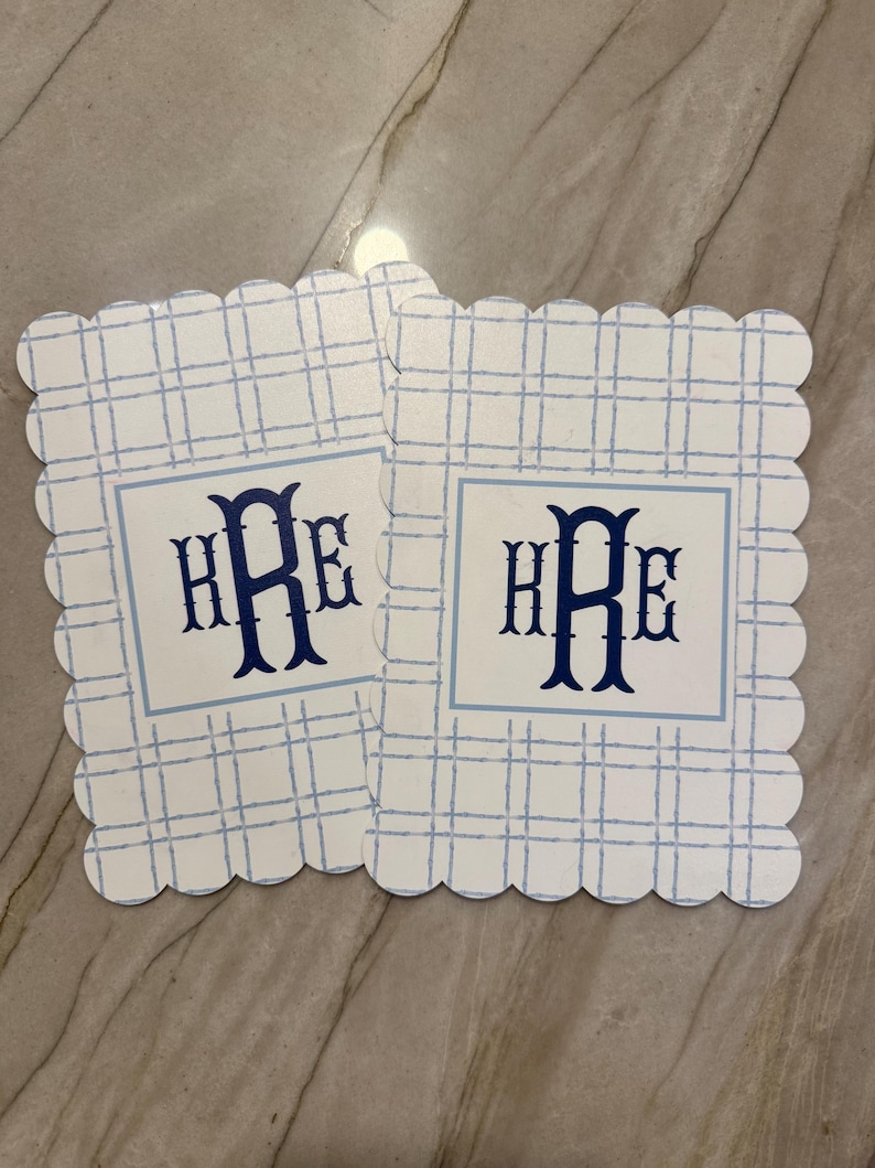 K&ouml;nnte beinhalten: Zwei quadratische Untersetzer mit gewelltem Rand. Jeder Untersetzer hat ein blau-wei&szlig;es Karomuster und ein zentrales Quadrat mit dem Monogramm "HRE" in Marineblau. Die Untersetzer sind aus einem robusten Material gefertigt.