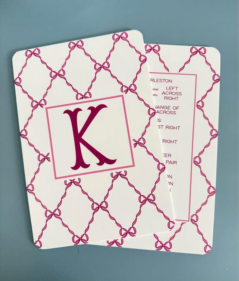 K&ouml;nnte beinhalten: Zwei wei&szlig;e Karten mit einem rosa Schleifenmuster und einem rosa Buchstaben "K" in einem rosa Quadrat. Eine Karte hat den Text "RLESTON" und andere W&ouml;rter. Die Karten liegen auf einer hellblauen Oberfl&auml;che.