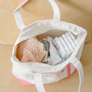 May include: A white canvas tote bag with pink accents and handles, featuring the name "Eliza" embroidered in pink on the front pocket. The open bag is filled with a soft peach-colored plush item, folded white fabric, and a stack of baby diapers.