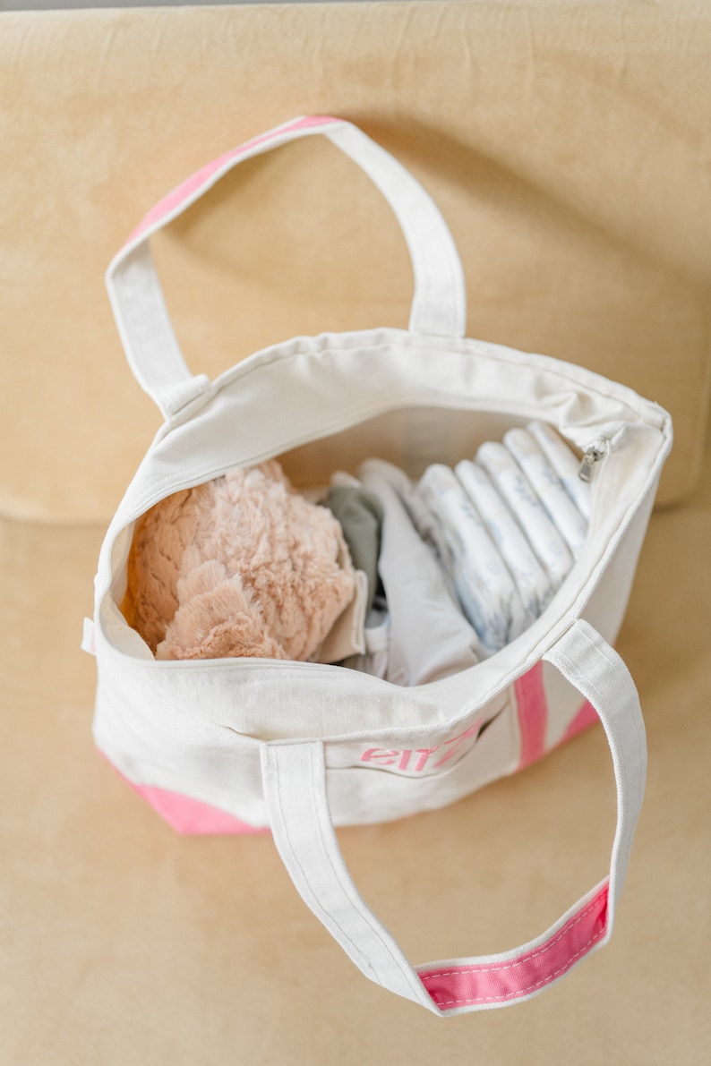 May include: A white canvas tote bag with pink accents and handles, featuring the name "Eliza" embroidered in pink on the front pocket. The open bag is filled with a soft peach-colored plush item, folded white fabric, and a stack of baby diapers.