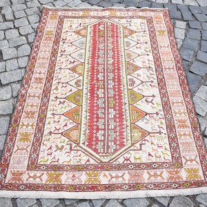 May include: A rectangular, cream-colored rug with a detailed geometric pattern. The rug features a central red stripe bordered by intricate designs in orange, green, and brown. Small animal figures are scattered across the cream field.